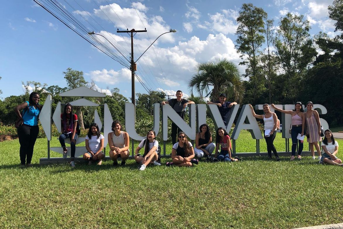 Imagem de intercambistas sentados em um gramado em frente a logo da Univates